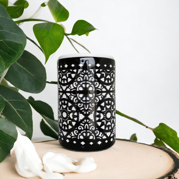 A black cylindrical diffuser or candle holder with a white geometric cutout pattern, displayed on a rustic wood slice pedestal with green leaves and white petals in the foreground.